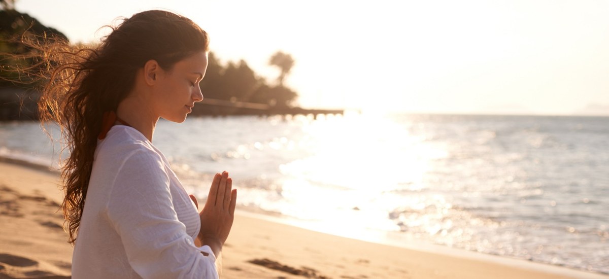 Femeie meditând pe plajă, cu palmele unite, la răsărit