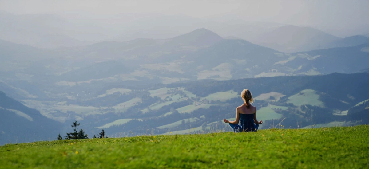 Persoană în meditație pe un deal verde, cu munții în depărtare