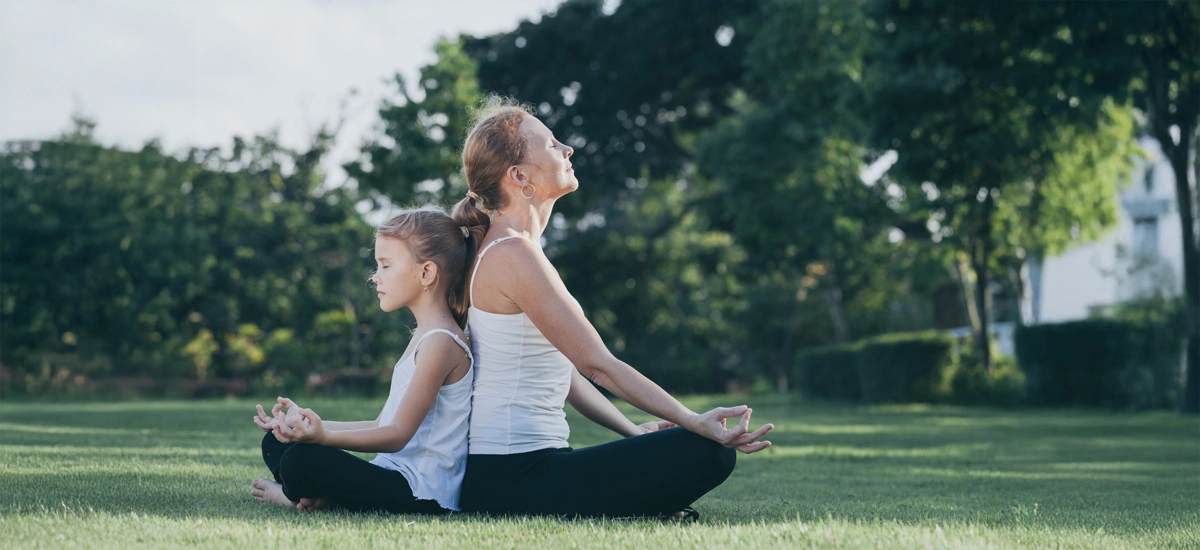 O profesoară și o fetiță așezate în meditație, una lângă alta, într-o grădină